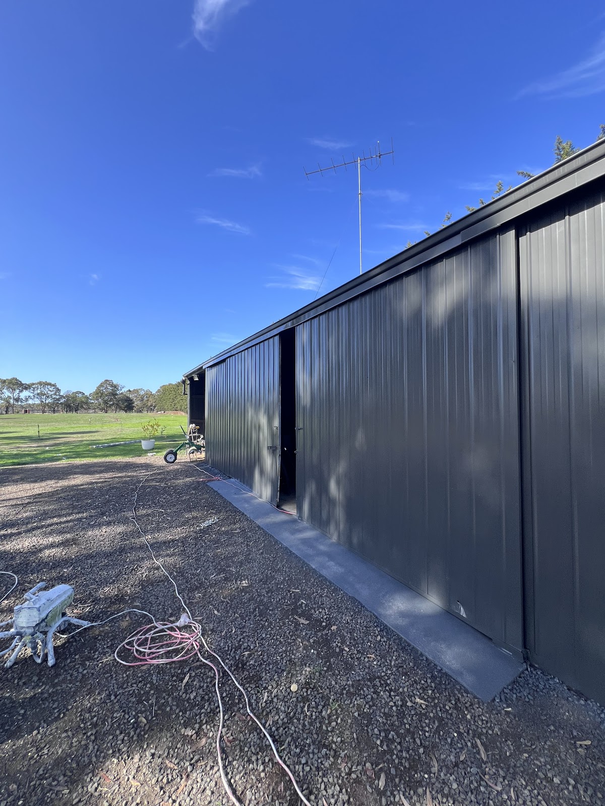 Long shed wall freshly painted dark charcoal, with painting gear on the gravel